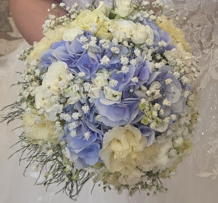 Bouquet tondo di ortensie azzurre, rose e lisianthus color crema e gypsophila davanti a un abito da sposa di pizzo