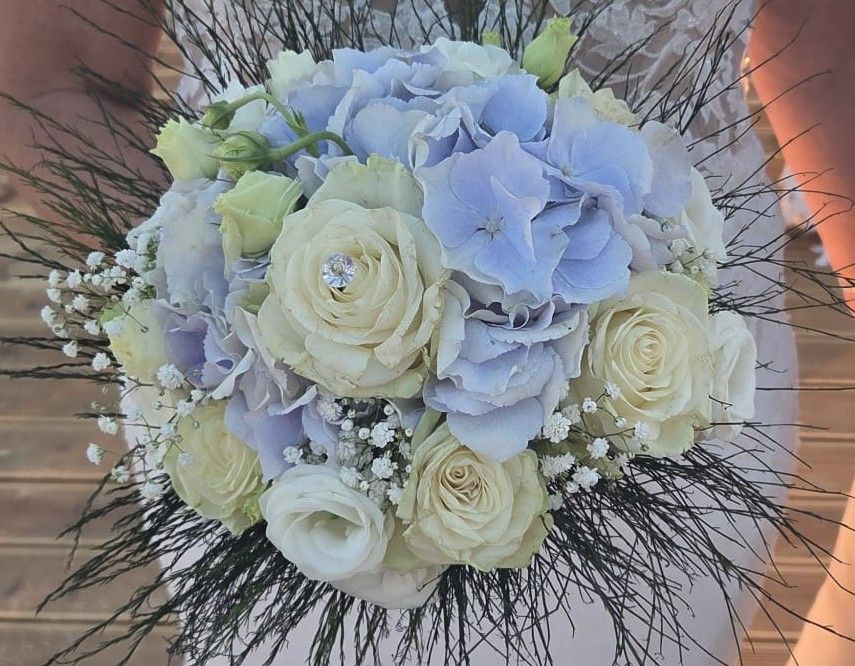 Bouquet tondo di ortensie azzurre, rose bianche, lisianthus e gypsophila con fili d'erba decorativi, tenuto dalla sposa