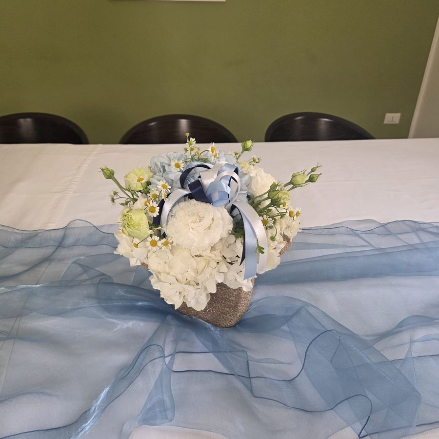 Composizione in cesto di juta con ortensie bianche e azzurre, garofani, lisianthus e camomilla, con fiocchi in raso azzurro e blu su tavolo con runner di organza celeste