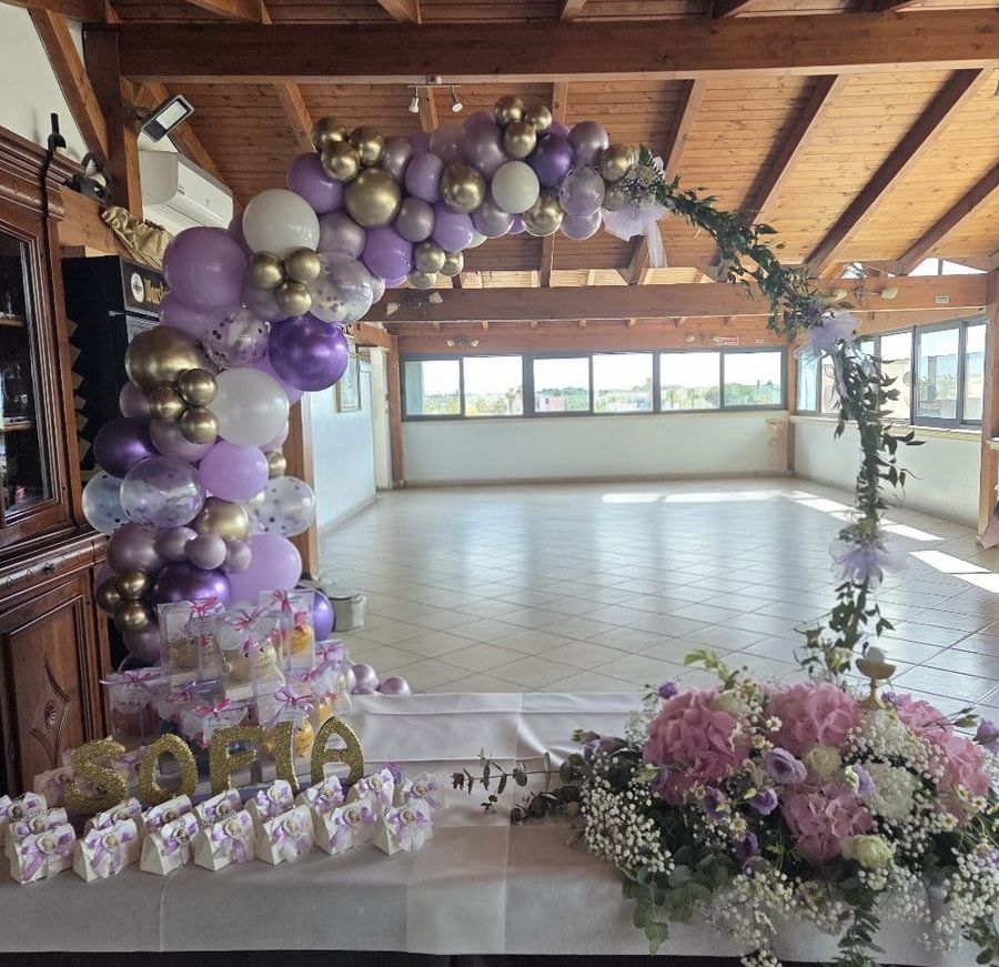 Sala per festa con travi in legno allestita con arco di palloncini lilla, argento e oro, nome della festeggiata in lettere dorate, bomboniere e composizione di ortensie rosa e gypsophila