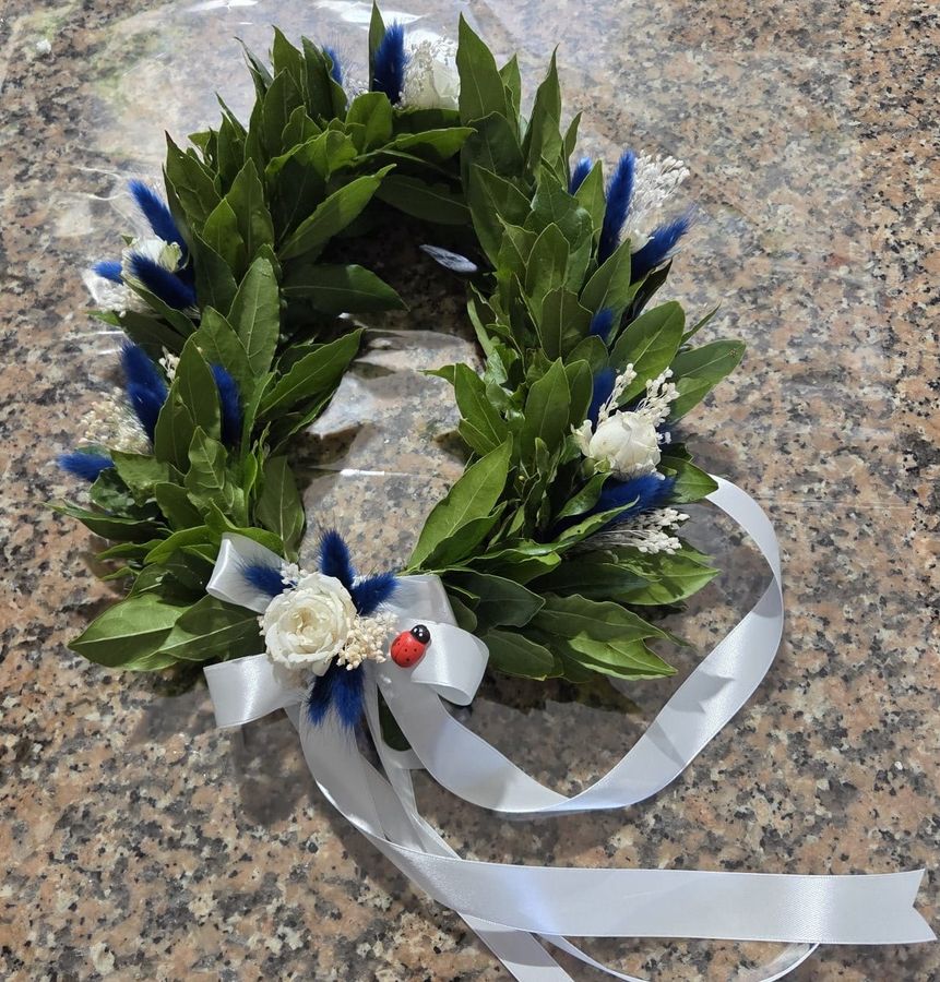 Corona d'alloro da laurea decorata con lagurus blu, roselline bianche stabilizzate, gypsophila, coccinella portafortuna e nastro di raso bianco