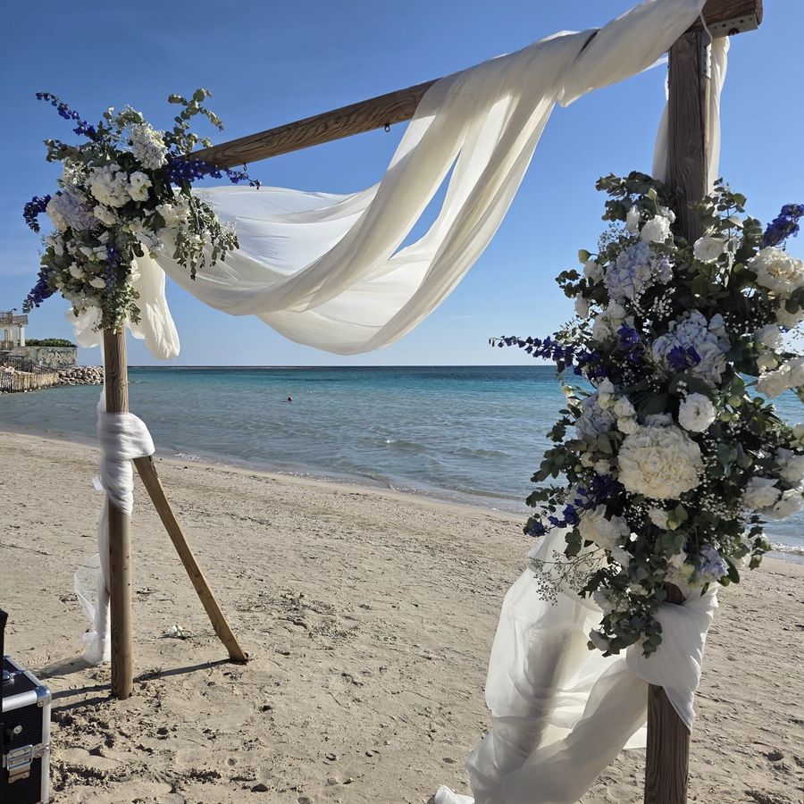 Arco di cerimonia in legno sulla spiaggia con teli di voile bianco al vento e composizioni di ortensie, rose bianche e delphinium blu