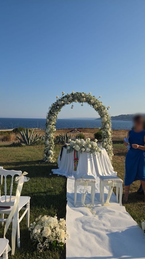 Arco floreale bianco per cerimonia civile sul mare con tavolo degli sposi addobbato e sedie bianche lungo il tappeto