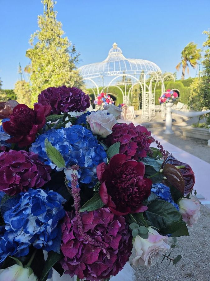 Composizione di ortensie blu e viola, peonie bordeaux e anthurium davanti al gazebo bianco di una cerimonia in giardino