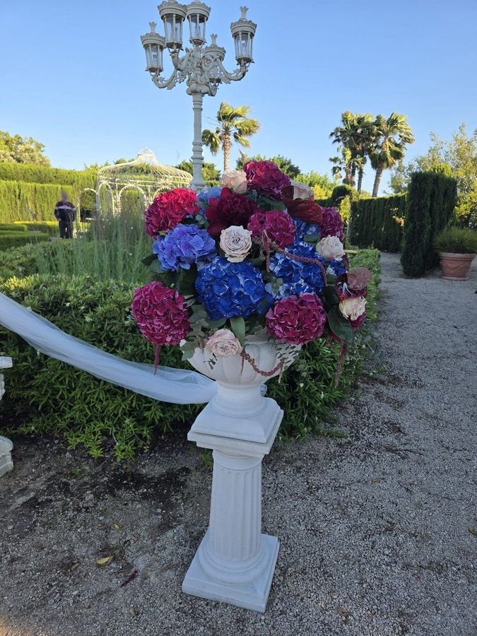 Grande composizione di ortensie blu e fucsia con rose in vaso classico bianco su colonna, all'ingresso del giardino della cerimonia