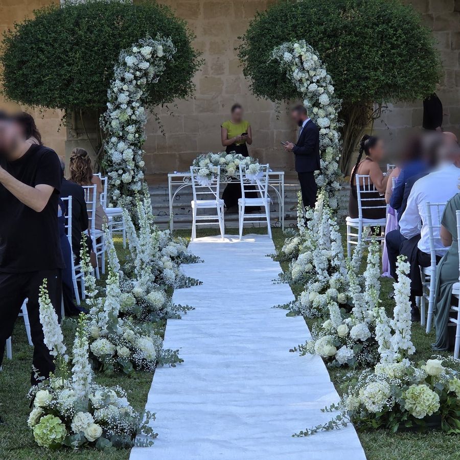 Allestimento di cerimonia all'aperto con tappeto bianco, arco floreale total white di rose, ortensie e gypsophila e composizioni a terra lungo la navata