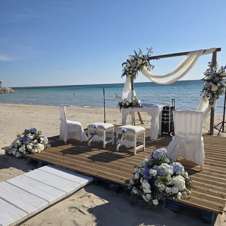 Allestimento di cerimonia civile in spiaggia con arco in legno drappeggiato di voile bianco e composizioni di ortensie azzurre e rose bianche