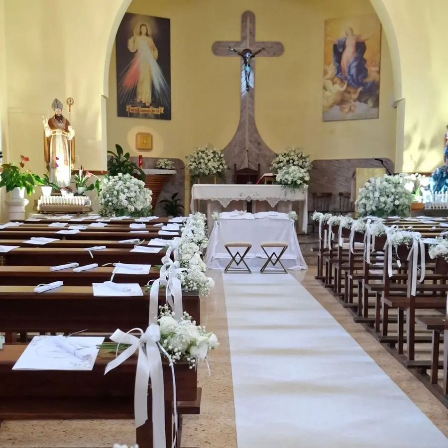 Navata di chiesa addobbata per matrimonio con bouquet bianchi sui banchi, tappeto nuziale e composizioni di rose bianche all'altare