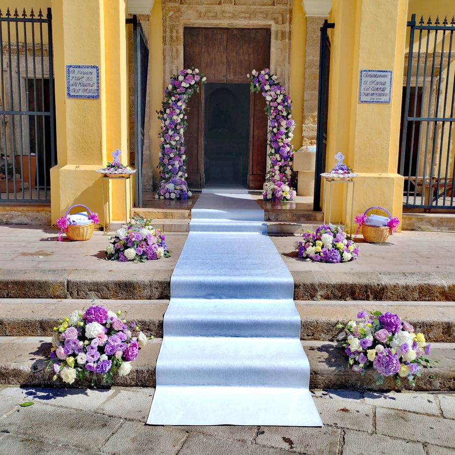 Ingresso di chiesa addobbato con arco floreale lilla e bianco, tappeto nuziale e composizioni a terra sui gradini