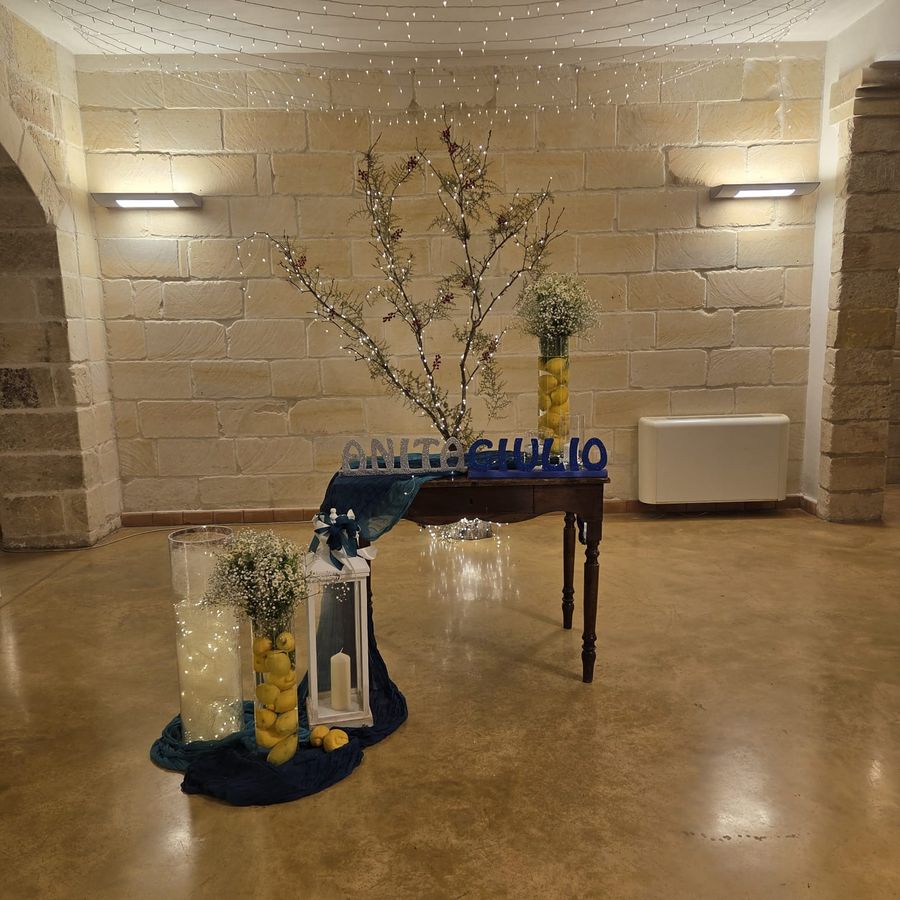 Angolo di benvenuto con scritte glitter dei nomi degli sposi, ramo luminoso, vasi con limoni, gypsophila e drappi blu su tavolo antico
