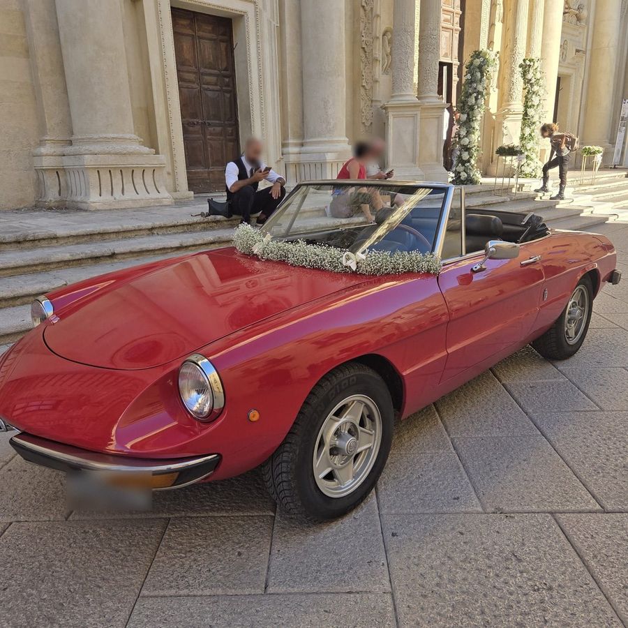 Auto d'epoca rossa decorata con ghirlanda di gypsophila e nastro bianco davanti al sagrato di una chiesa barocca