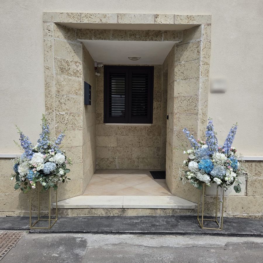 Coppia di composizioni floreali su alzate dorate con ortensie azzurre, delphinium, lisianthus e nebbiolina ai lati di un portone in pietra leccese