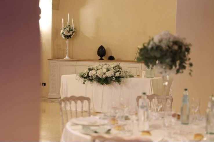 Sala ricevimento con tavolo degli sposi addobbato di fiori bianchi e centrotavola alti su tavoli rotondi apparecchiati