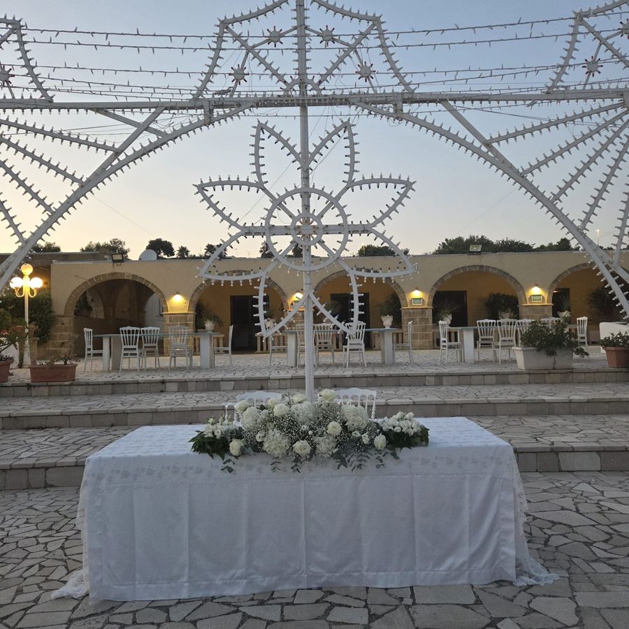 Tavolo degli sposi allestito all'aperto con lunga composizione di rose bianche, ortensie e gypsophila sotto le luminarie salentine