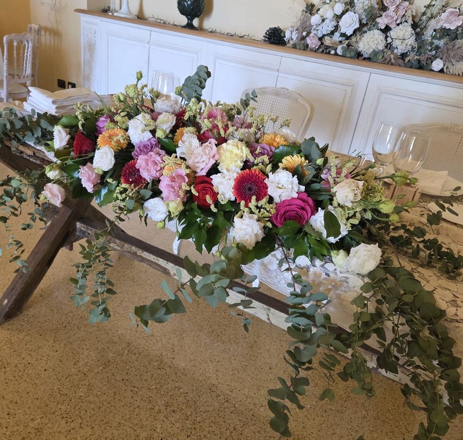 Lunga composizione di rose, gerbere, garofani e lisianthus nei toni del rosa, del rosso e del bianco con eucalipto ricadente, su un supporto rustico in legno accanto al tavolo