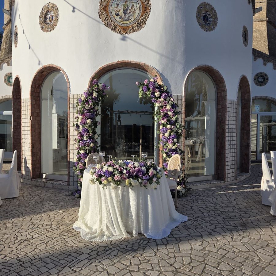 Tavolo degli sposi con tovaglia in pizzo avorio e runner floreale lilla, incorniciato da un arco di rose e ortensie viola e bianche davanti alla masseria