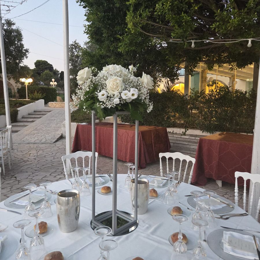 Centrotavola alto su struttura metallica con rose bianche, lisianthus e gypsophila su tavolo di ricevimento all'aperto al tramonto