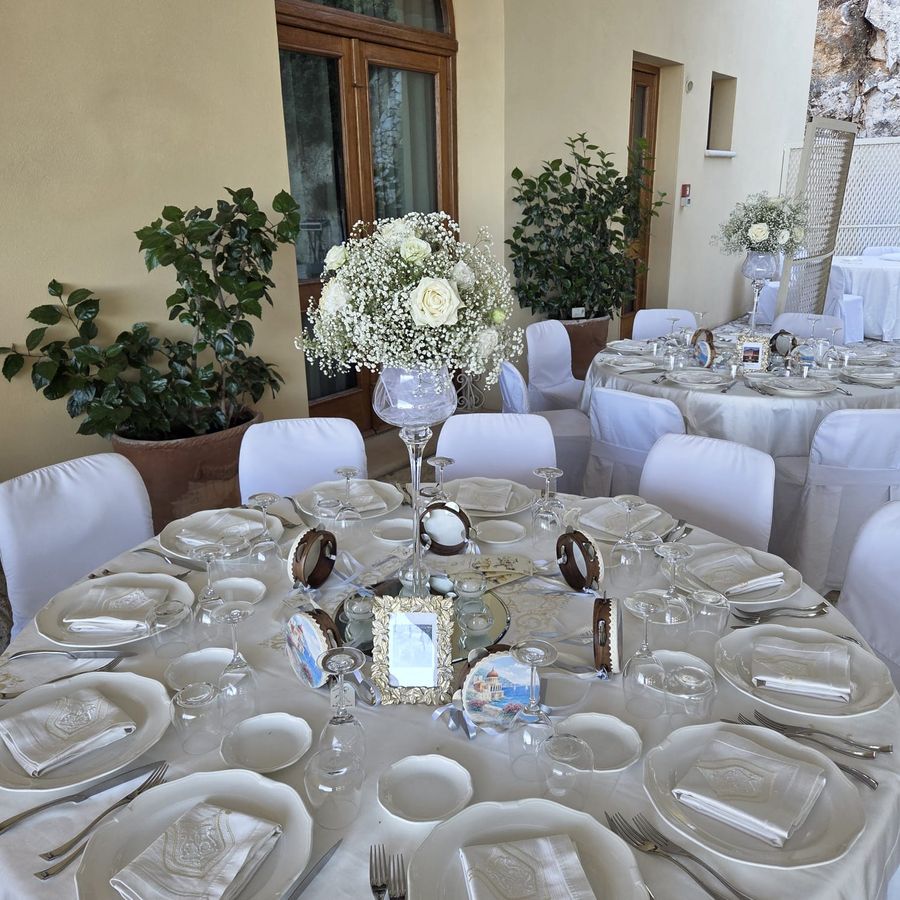 Tavolo di ricevimento apparecchiato in bianco con centrotavola alto in coppa di vetro di rose bianche e nebbiolina