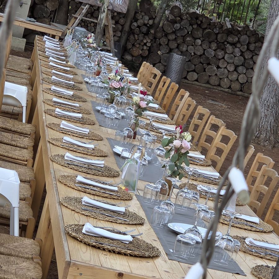 Tavolo imperiale allestito all'aperto in pineta con mise en place completa, tovagliette in rafia e piccoli vasi con rose rosa ed eucalipto